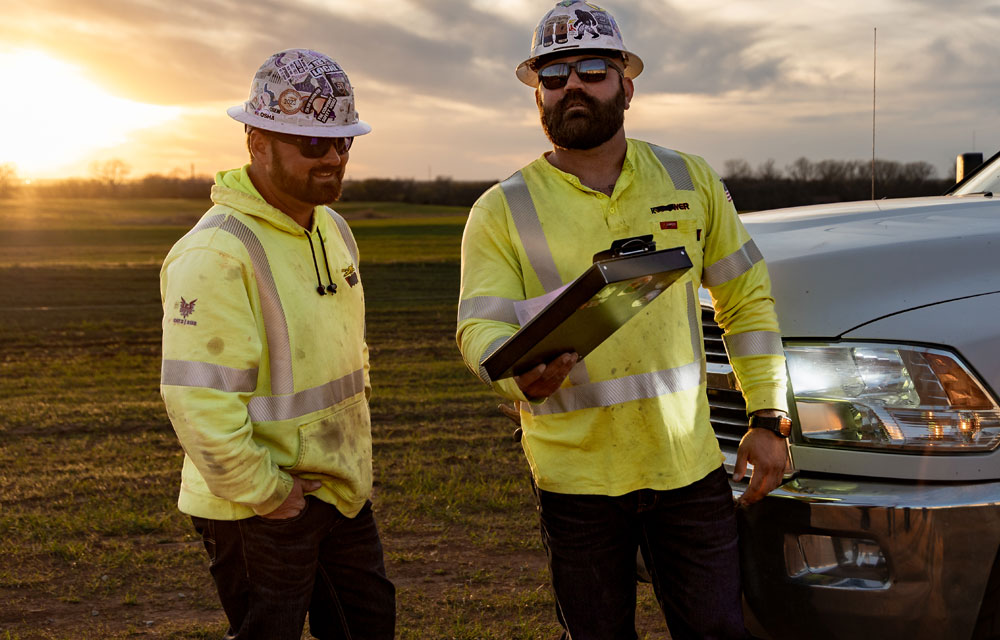 Two electrical linemen, Dylan Edgmon and Kevin Walker, in high-visibility jackets and hard hats stand beside a work truck at sunset, one holding a clipboard as they review a task.